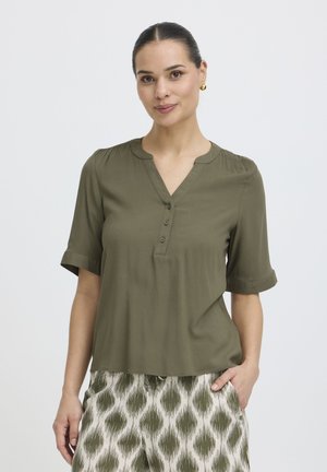 Kobieta ubrana w oliwkowo-zieloną bluzkę z krótkim rękawem i guzikami oraz wzorzyste beżowo-zielone spodnie, stojąca z jedną ręką w kieszeni.