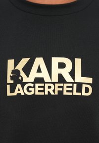 Čierna tkanina s béžovým textom "KARL LAGERFELD" a siluetou muža s okuliarmi a konským chvostom vo vnútri písmena K.