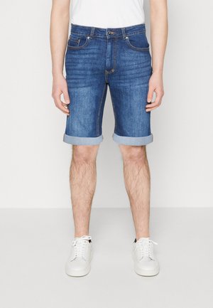 Männliches Model trägt hochgekrempelte blaue Jeansshorts, weiße Turnschuhe und ein weißes T-Shirt, steht vor einem schlichten hellen Hintergrund.
