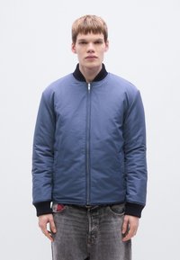 Μπλε μπουφάν bomber με φερμουάρ μπροστά, ριμπ γιακά και μανσέτες. Συνδυάζεται με γκρι τζιν παντελόνι, φορεμένο με τα χέρια στις τσέπες.