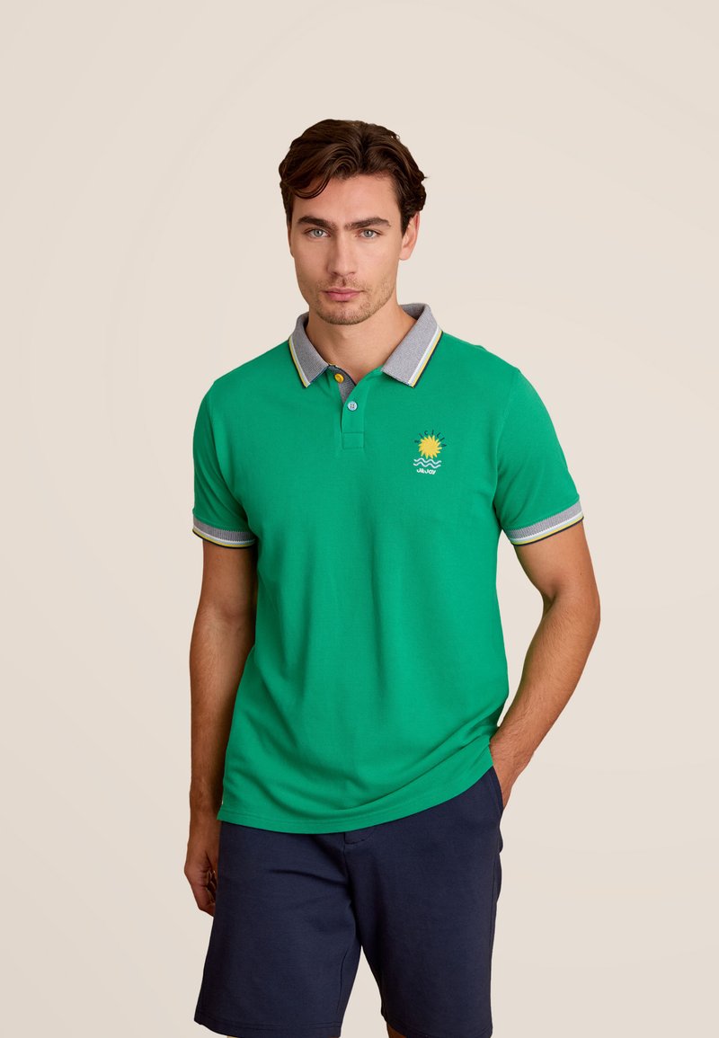 Groene poloshirt met grijze kraag en mouwenaccenten, voorzien van een geel zonlogo op de borst. Gepaard met marineblauwe shorts.