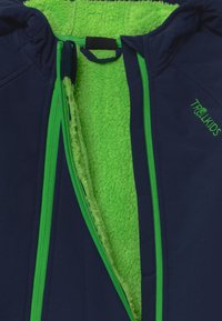 En marinblå jacka med en ljusgrön fleecefoder, med gröna dragkedjor och logotyp. Insidan har en texturerad, fluffig yta.