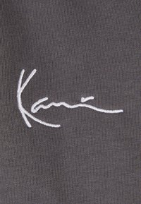 Tejido gris con un diseño de firma bordada en blanco, que presenta curvas suaves y un estilo de escritura fluido. Textura detallada del tejido visible.