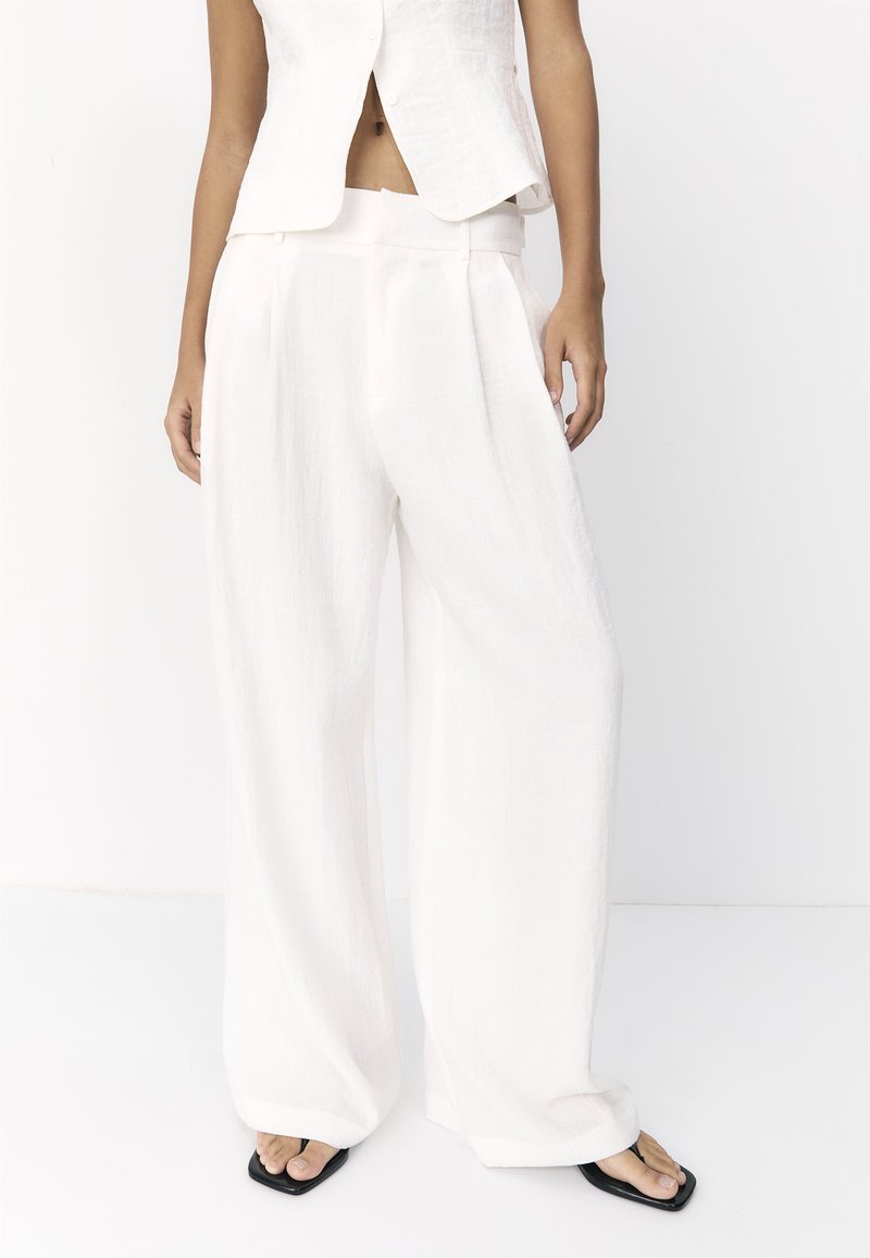Personne portant un pantalon blanc à jambes larges, un gilet blanc sans manches et des sandales noires à bout carré, debout devant un fond blanc uni.