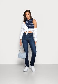 Top sem mangas às riscas azul marinho, camisa branca atada à cintura, jeans azul escuro, mala azul clara e ténis brancos. Textura suave, estilo casual.