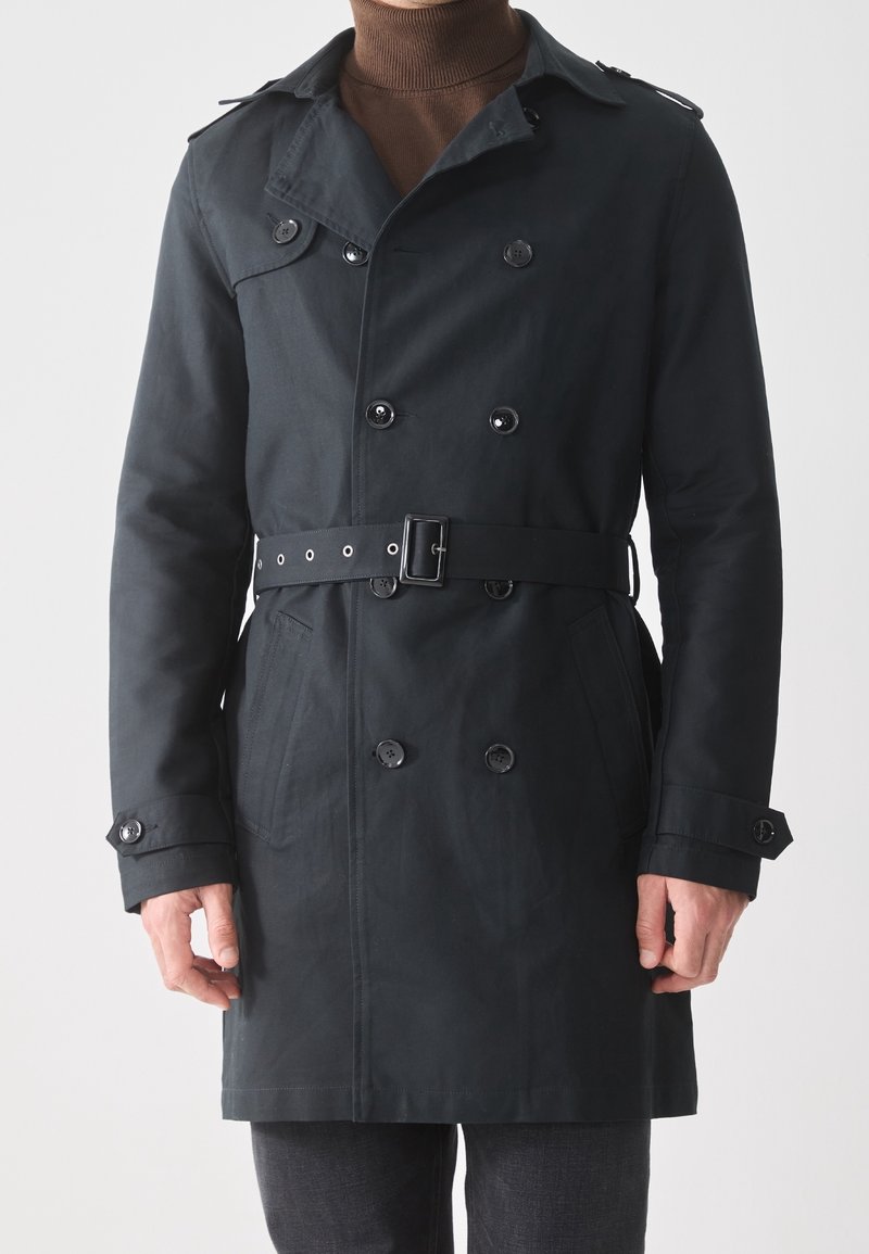 Homme portant un trench-coat noir croisé à ceinture à la taille et poignets boutonnés sur un col roulé marron.