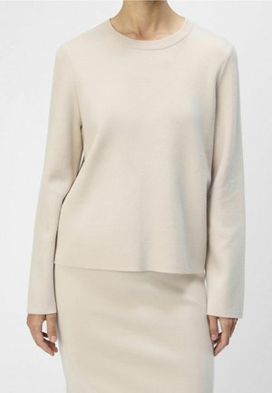 Kvinde iført beige langærmet striktop og matchende nederdel, stående mod en ensfarvet lys baggrund.