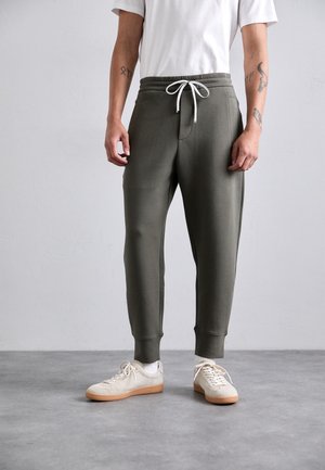 Man in olijfgroene joggerbroek met witte trekkoord, witte sokken, beige sneakers en een wit shirt met korte mouwen, staande op een grijze vloer.