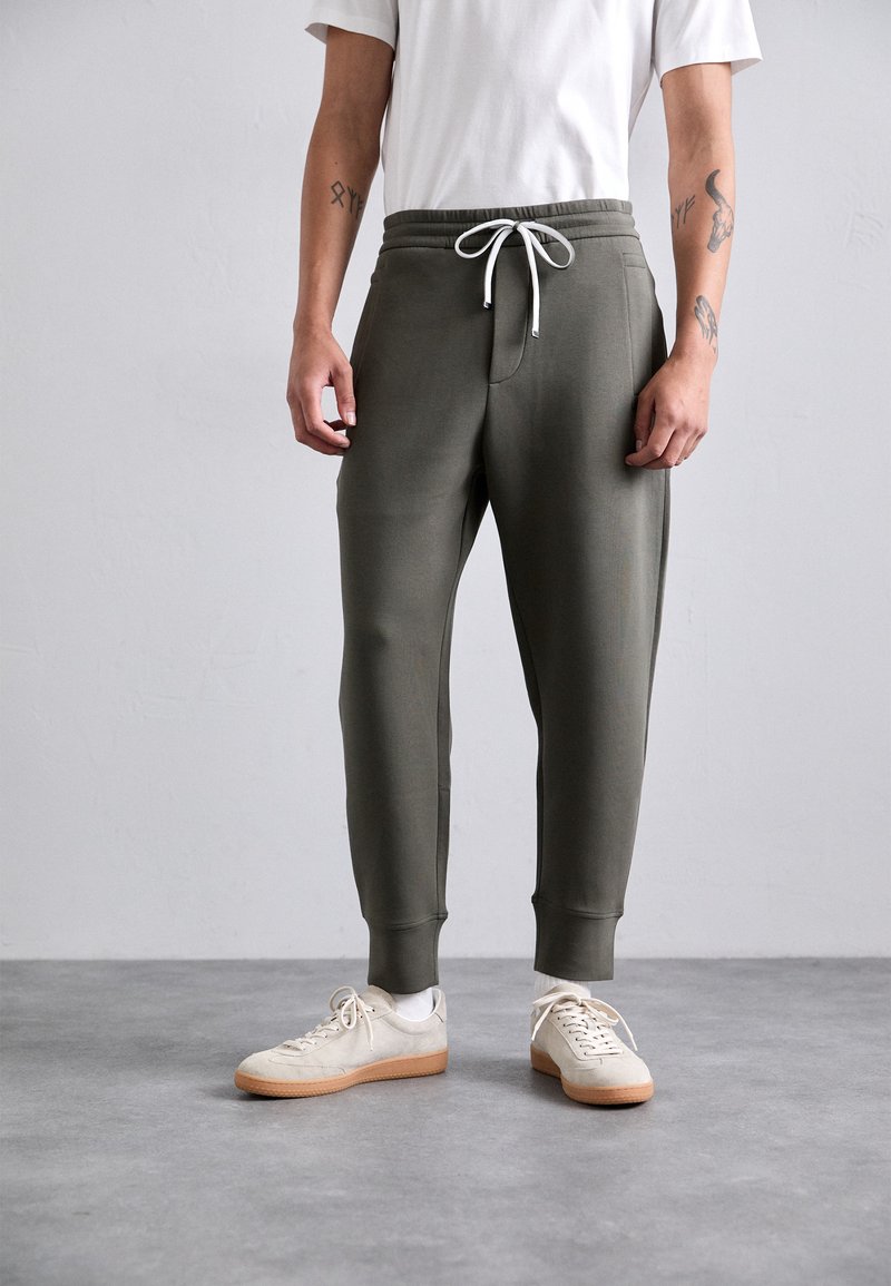 Mann iført olivengrønne joggebukser med hvit snor, hvite sokker, beige sneakers og en hvit kortermet skjorte, stående på grått gulv.