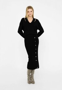 Pulover negru tricotat cu decolteu în V, asortat cu o fustă lungă neagră cu detalii cu nasturi. Accesorii includ cizme înalte până la genunchi cu model.