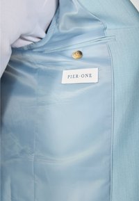 Interno di giacca azzurra chiara con texture liscia, dotato di un'etichetta bianca contrassegnata "PIER·ONE" e un unico bottone dorato.