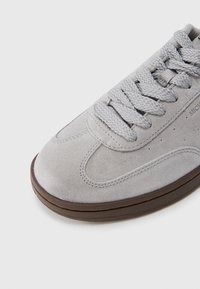 Ανοιχτό γκρι σουέντ αθλητικό παπούτσι με παχιά υφασμάτινα κορδόνια και σκούρα καφέ σόλα, εμφανίζεται από μπροστινή επάνω γωνία σε λευκό φόντο.