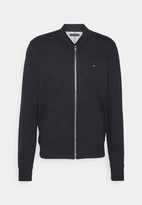 Chaqueta bomber negra con cremallera, cuello, puños y dobladillo de canalé, bolsillos frontales y un pequeño logo en rojo, blanco y azul en el pecho.