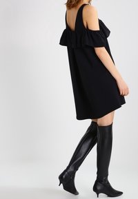 Woman wearing a black off-shoulder mini dress with ruffles and knee-high black leather heeled boots, standing against a plain background.