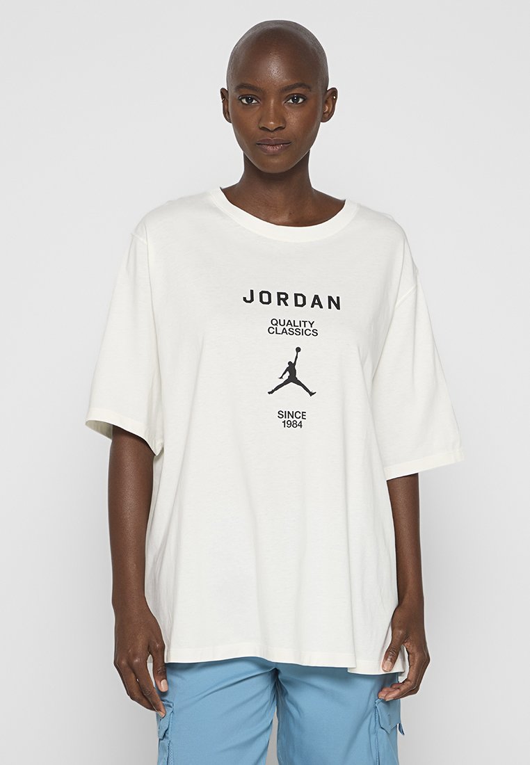 Osoba ubrana w białą, oversize'ową koszulkę Jordan z czarnym logo i napisem, w połączeniu z niebieskimi spodniami cargo, stojąca na tle jednolitym.