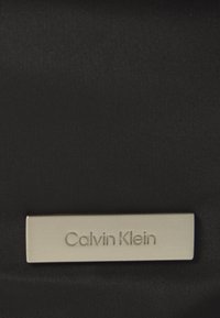 Zwarte stof met een gladde textuur, voorzien van een zilveren rechthoekig plaquette waarin de tekst "Calvin Klein" is gegraveerd.