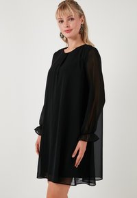 Black dress made of lightweight fabric with long sheer sleeves, a round neckline, and ruffled cuffs. The silhouette is loose and knee-length.