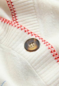White wool fabric with ribbed texture and red stitching detail. Features a dark brown button with four holes. Close-up perspective.