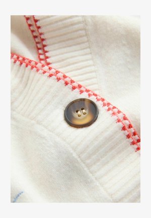 Weißer Wollstoff mit gerippter Textur und roten Nähten. Verfügt über einen dunkelbraunen Knopf mit vier Löchern. Nahaufnahme.