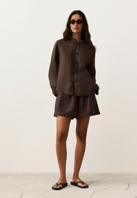 Femme portant une veste marron foncé boutonnée et un short assorti avec des sandales noires et des lunettes de soleil, debout sur un sol carrelé devant un fond uni.