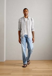 Cămașă cu nasturi, cu dungi verticale alb-negru, tricou alb, blugi de culoare albastră deschisă și sandale maro, pe un fundal neutru.