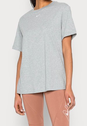 Vrouw die een lichtgrijs kortemouwen Nike T-shirt en lichtbruine legging met wit Nike-logo op de dij draagt, staand tegen een effen achtergrond.