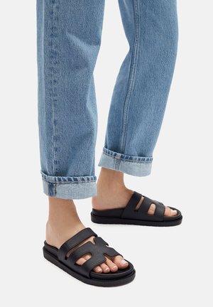 Czarne klapki na slajdzie z trzema wycięciami na paskach, teksturowana gumowa podeszwa, zestawione z podwiniętymi jasnoniebieskimi jeansami.