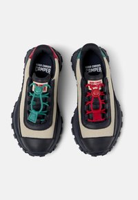Zapatillas de deporte en tonos marrones y negros con cordones verdes y rojos, suelas de goma y lados de malla textil. Presentan un diseño voluminoso con acentos de color distintivos.