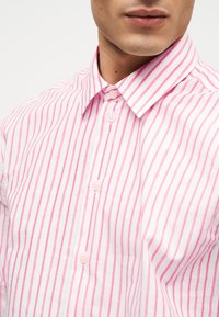 Άνδρας που φοράει ροζ και λευκό πουκάμισο με κάθετες ρίγες και κουμπιά, με μυτερό γιακά, απεικονίζεται από τον λαιμό έως το στήθος.