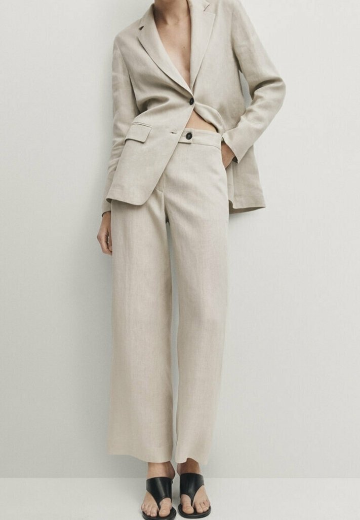 Blazer en lin beige à un bouton, assorti à un pantalon large et des sandales à talons noires à bout ouvert, modèle avec une main dans la poche.