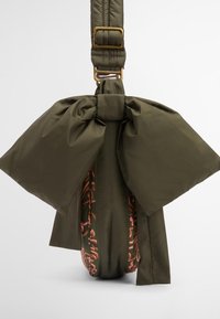Zielona oliwkowa torebka z dużą kokardą, z teksturowanym wykończeniem oraz wzorzystym wnętrzem w odcieniach różu. Regulowany pasek z złotymi akcentami.