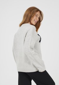 Uśmiechnięta kobieta z długimi rudymi włosami, ubrana w jasnoszary sweter dzianinowy i czarne spodnie, pozująca bokiem na białym tle.