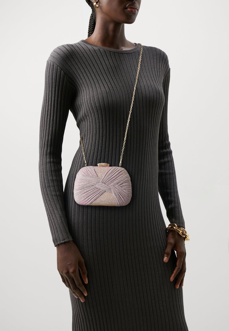 Borsa a tracolla di perline rosa con un design incrociato testurizzato e una catena, tenuta contro un vestito grigio a coste.