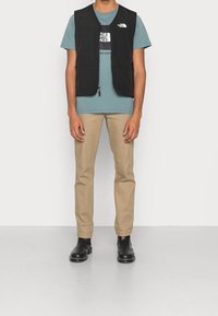 Czarna bezrękawnik z dwoma przednimi kieszeniami, noszony na turkusowej koszulce z logo, w połączeniu z beżowymi spodniami i czarnymi botkami za kostkę.