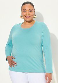 Ein langärmliges, türkisfarbenes Oberteil aus glattem Stoff mit Rundhalsauschnitt, das eine lockere Passform und dezenten Stretch aufweist.