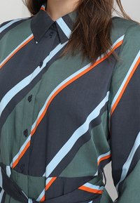 Napapaita, joka on tummanvihreää kangasta ja jossa on vinoja raitoja sinisessä, oranssissa ja valkoisessa. Sisältää kauluksen ja sidontalan yksityiskohdan vyötäröllä.