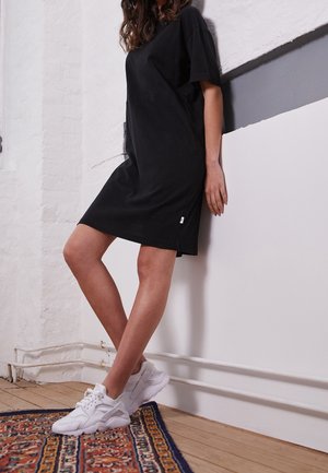 Femme appuyée contre un mur blanc, portant une robe noire et des baskets blanches, debout sur un tapis à motifs sur un sol en bois.