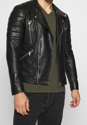 Homme portant une veste en cuir matelassée noire avec des fermetures éclair argentées sur un t-shirt vert olive et un jean noir.