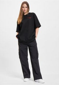 Black oversized t-shirt with red logo, paired with loose black cargo pants featuring side pockets, worn with white sneakers.