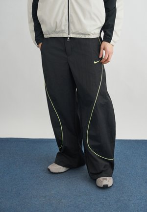 Asmuo, dėvintis juodas „Nike“ sportines kelnes su neoninėmis siūlėmis, šviesiai pilkus sportinius batelius ir baltai juodą striukę, stovintis ant mėlyno kilimo.