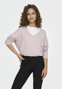 Pull en V oversize rose clair superposé à un t-shirt blanc à col rond, associé à un jean noir ajusté. Tissu lisse, coupe décontractée.