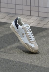Baskets en cuir blanc avec des accents en suède beige, un onglet de talon bleu marine et un logo doré. Semelle texturée avec un motif décoratif. Design classique à lacets.