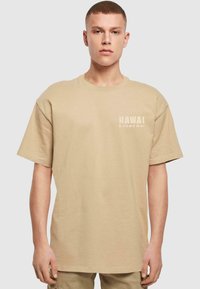 Beige bomulds t-shirt med korte ærmer, rund hals og trykt tekst "HAWAI E KOMO MAI" i en lys, diskret farve på venstre bryst.
