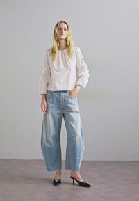 Fehér, puffos ujjú blúz, gyűrött nyakkal, világoskék, bő szárú farmernadrággal és fekete, hegyes orrú magassarkú cipővel egy semleges háttér előtt.