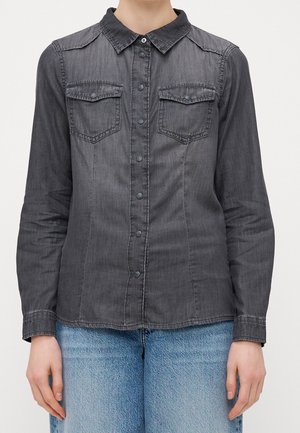 Mørkegrå denimskjorte med knapper og to brystlommer, båret med lyseblå jeans, vist fra skuldre til hofter.