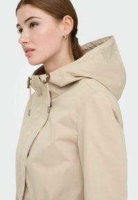 Veste imperméable beige avec capuche, présentant une texture lisse, des fermetures à cordon de serrage et un design minimaliste sans motifs visibles.