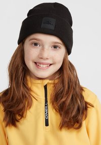 Black knit beanie with a rectangular label featuring a logo. The wearer is in a yellow fleece top with a half-zip collar and black accents.