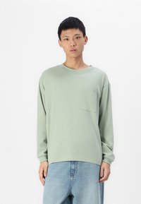 Lange mouw, lichtgroen katoenen t-shirt met een ronde hals en een voorzak, gecombineerd met loszittende lichtblauwe jeans.