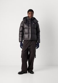 Juodas pūkinis švarkas su aukštu apykakle, blizgiu paviršiumi ir užtrauktuku, derinamas su laisvomis krovinių kelnėmis ir mėlynomis pirštinėmis. Tvirti juodi batai.