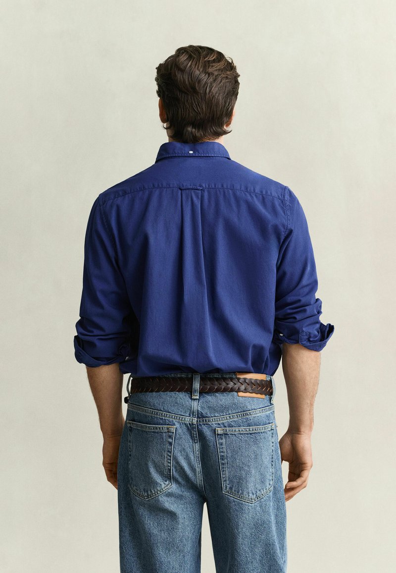 Mann, der mit dem Rücken zur Kamera steht, trägt ein blaues Hemd mit langen Ärmeln, die hochgekrempelt sind, und hellblaue Jeans mit braunem Gürtel.